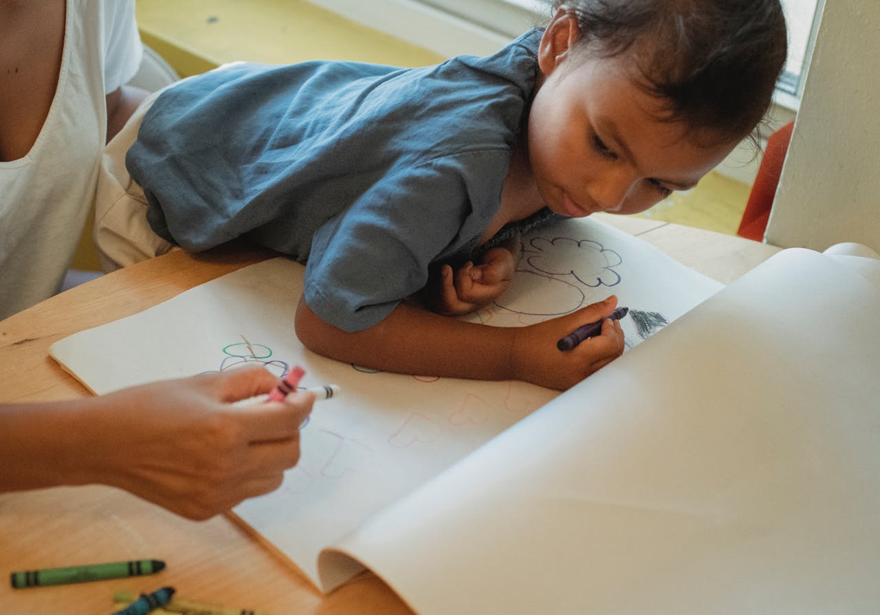 Crop anonymous female drawing with color wax crayons together with concentrated little daughter at table