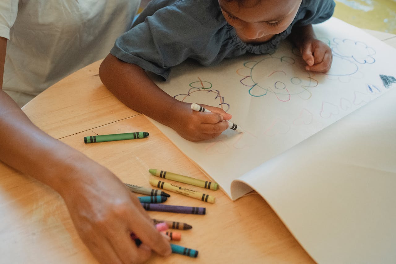 From above of crop little ethnic girl drawing in album with colorful crayons while sitting on knees of unrecognizable mother at home
