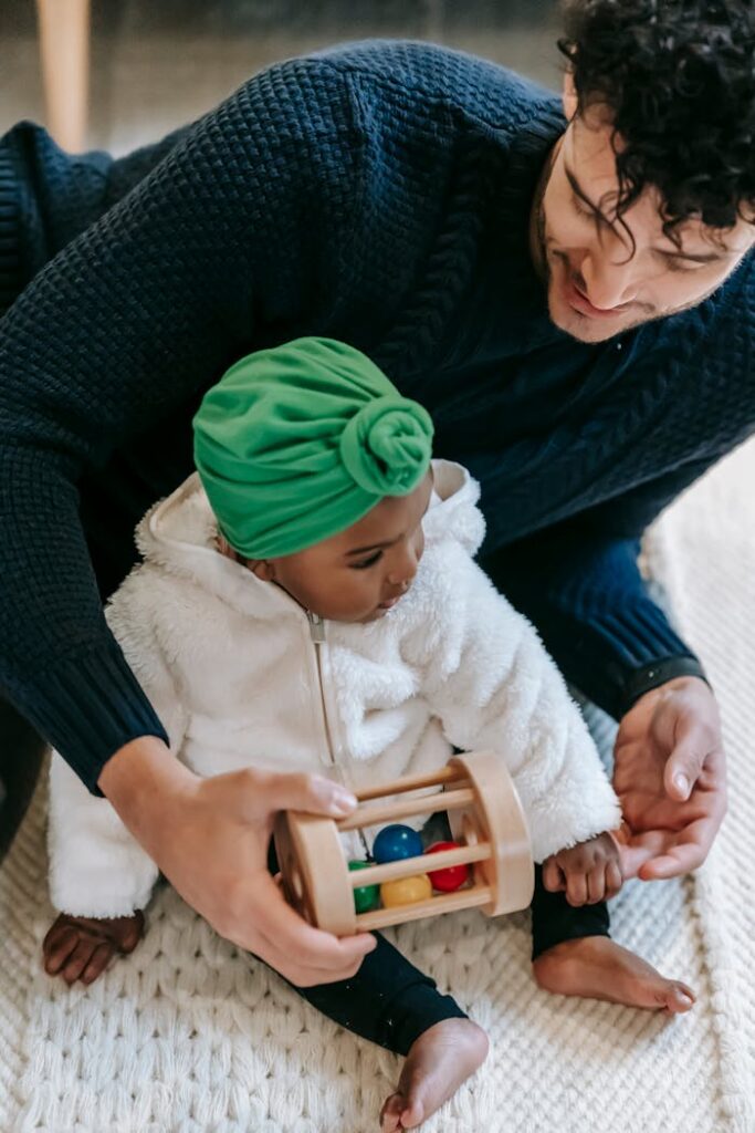 From above of crop happy ethnic man in casual clothes playing with cute black daughter in turban sitting on carpet at home