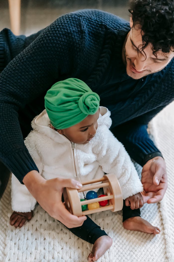 From above of crop happy ethnic man in casual clothes playing with cute black daughter in turban sitting on carpet at home